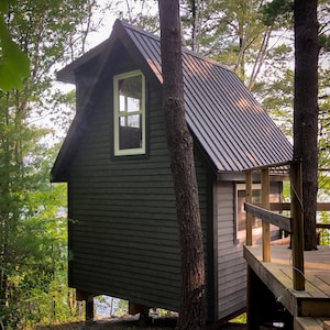 May include: A small, dark gray cabin with a metal roof and a single window. The cabin is built on stilts and is surrounded by trees.
