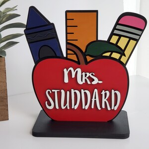K&ouml;nnte beinhalten: Ein roter Apfel mit einem gr&uuml;nen Blatt, einem blauen Buntstift, einem gelben Bleistift mit einem rosa Radiergummi und einem orangefarbenen Lineal. Der Apfel tr&auml;gt den Text "Mrs. Studdard".
