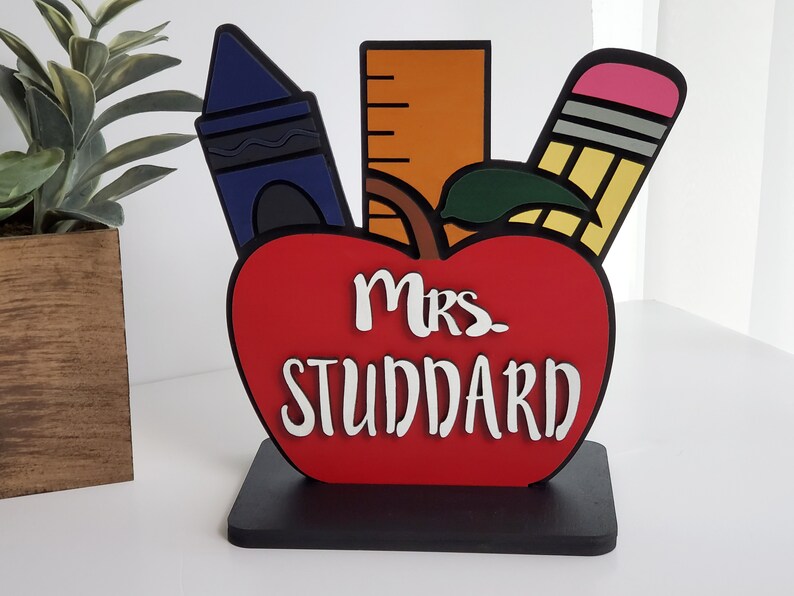 K&ouml;nnte beinhalten: Ein roter Apfel mit einem gr&uuml;nen Blatt, einem blauen Buntstift, einem gelben Bleistift mit einem rosa Radiergummi und einem orangefarbenen Lineal. Der Apfel tr&auml;gt den Text "Mrs. Studdard".