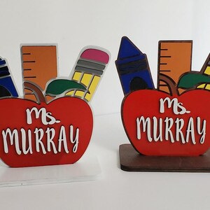 K&ouml;nnte beinhalten: Zwei rote, apfelartige Holzschilder f&uuml;r Lehrertische mit einem Lineal, einem Buntstift und einem blauen Buntstift. Die Schilder zeigen "Ms. Murray" in wei&szlig;en Buchstaben.