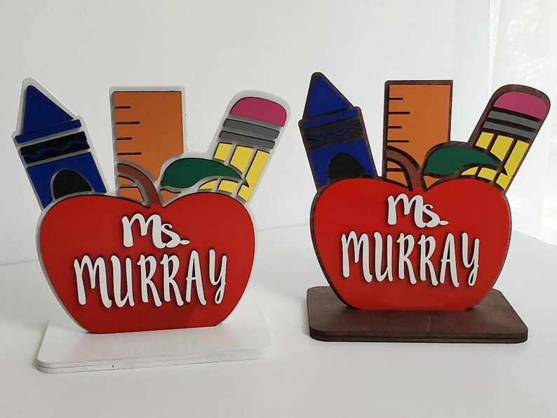K&ouml;nnte beinhalten: Zwei rote, apfelartige Holzschilder f&uuml;r Lehrertische mit einem Lineal, einem Buntstift und einem blauen Buntstift. Die Schilder zeigen "Ms. Murray" in wei&szlig;en Buchstaben.