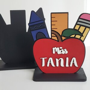 K&ouml;nnte beinhalten: Ein roter Apfel mit einem schwarzen Stiel und einem gr&uuml;nen Blatt, aus dem ein Lineal, ein Buntstift und ein Bleistift herausragen. Der Apfel tr&auml;gt die Aufschrift "Miss Tania" in wei&szlig;.