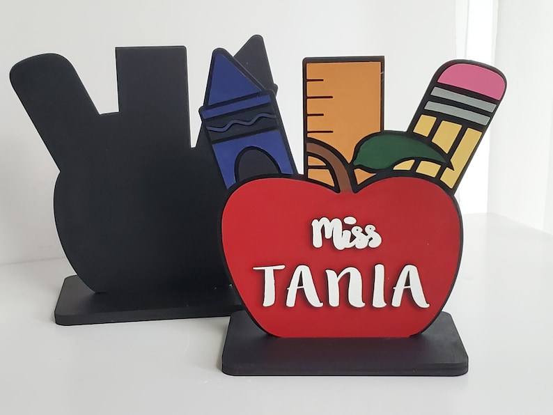 K&ouml;nnte beinhalten: Ein roter Apfel mit einem schwarzen Stiel und einem gr&uuml;nen Blatt, aus dem ein Lineal, ein Buntstift und ein Bleistift herausragen. Der Apfel tr&auml;gt die Aufschrift "Miss Tania" in wei&szlig;.