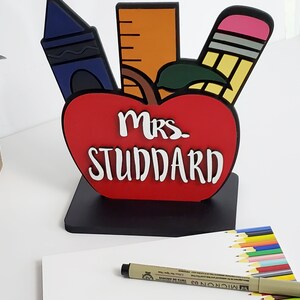 K&ouml;nnte beinhalten: Ein roter, apfelf&ouml;rmiger, h&ouml;lzerner Schreibtisch-Organizer mit schwarzem Sockel. Der Apfel hat ein gr&uuml;nes Blatt und ist mit einem Lineal, einem Buntstift und einem Bleistift verziert. Der Text "Mrs. Studdard" ist in Wei&szlig; auf den Apfel geschrieben.