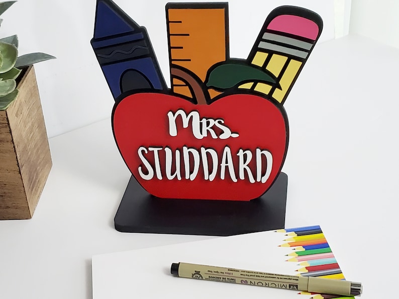 K&ouml;nnte beinhalten: Ein roter, apfelf&ouml;rmiger, h&ouml;lzerner Schreibtisch-Organizer mit schwarzem Sockel. Der Apfel hat ein gr&uuml;nes Blatt und ist mit einem Lineal, einem Buntstift und einem Bleistift verziert. Der Text "Mrs. Studdard" ist in Wei&szlig; auf den Apfel geschrieben.