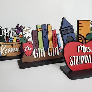 K&ouml;nnte beinhalten: Drei h&ouml;lzerne Lehrertischschilder mit bunten Illustrationen. Das erste Schild sagt "Create" und hat einen Korb mit Schulutensilien. Das zweite Schild sagt "Ms. Gin Gin" und hat einen Stapel B&uuml;cher. Das dritte Schild sagt "Mrs. Studdard" und hat einen roten Apfel.