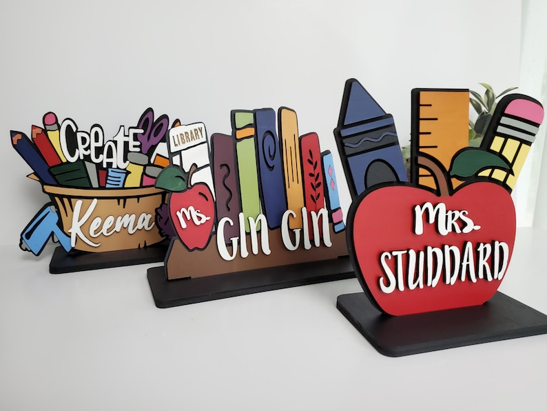 K&ouml;nnte beinhalten: Drei h&ouml;lzerne Lehrertischschilder mit bunten Illustrationen. Das erste Schild sagt "Create" und hat einen Korb mit Schulutensilien. Das zweite Schild sagt "Ms. Gin Gin" und hat einen Stapel B&uuml;cher. Das dritte Schild sagt "Mrs. Studdard" und hat einen roten Apfel.