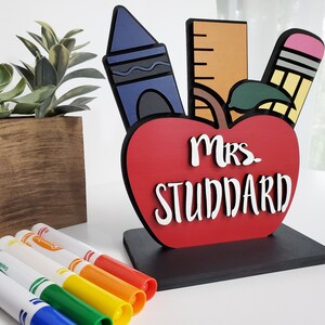 K&ouml;nnte beinhalten: Ein rotes, apfelf&ouml;rmiges Holzschild mit dem Text "Mrs. Studdard" in wei&szlig;en Buchstaben. Der Apfel ist mit einem blauen Buntstift, einem gelben Bleistift und einem Lineal verziert. Das Schild steht auf einem schwarzen Sockel.