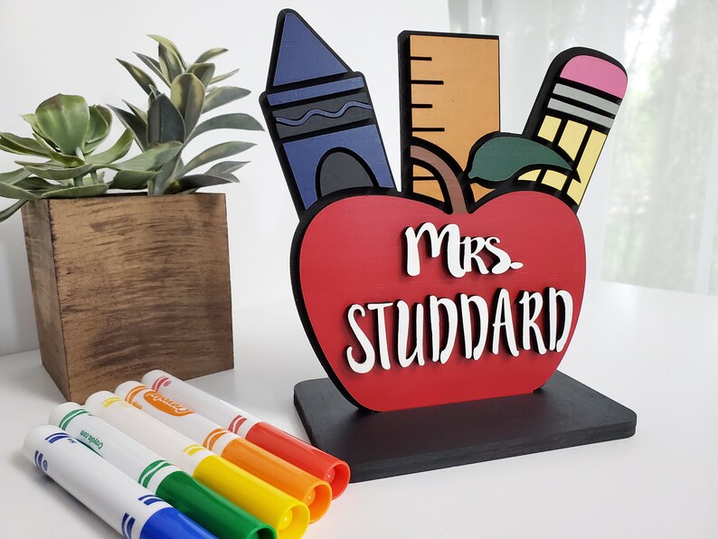 K&ouml;nnte beinhalten: Ein rotes, apfelf&ouml;rmiges Holzschild mit dem Text "Mrs. Studdard" in wei&szlig;en Buchstaben. Der Apfel ist mit einem blauen Buntstift, einem gelben Bleistift und einem Lineal verziert. Das Schild steht auf einem schwarzen Sockel.