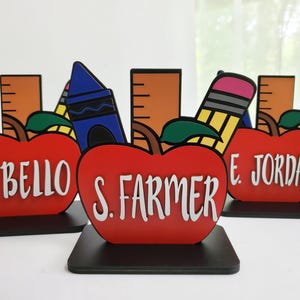 K&ouml;nnte beinhalten: Drei rote, apfelartige Lehrer-Dankes-Auszeichnungen mit schwarzen Sockeln. Jeder Apfel tr&auml;gt den Namen eines anderen Lehrers: R. Bello, S. Farmer und E. Jordan. Jeder Apfel ist mit einem Buntstift, einem Lineal und einem Bleistift verziert.