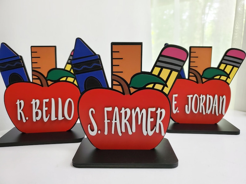 K&ouml;nnte beinhalten: Drei rote, apfelartige Lehrer-Dankes-Auszeichnungen mit schwarzen Sockeln. Jeder Apfel tr&auml;gt den Namen eines anderen Lehrers: R. Bello, S. Farmer und E. Jordan. Jeder Apfel ist mit einem Buntstift, einem Lineal und einem Bleistift verziert.