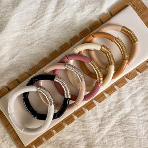 May include: A collection of six colorful bracelets. The bracelets are in shades of white, black, pink, and tan, with gold and silver wire accents. The bracelets are arranged on a white surface with a wooden border.