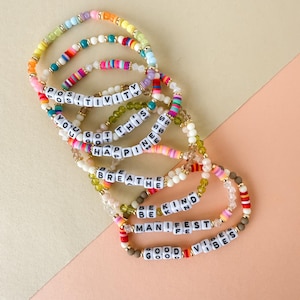 May include: A collection of colorful beaded bracelets. Each bracelet features a different positive message spelled out in white letter beads, including "POSITIVITY", "YOU GOT THIS", "BREATHE", "BE KIND", "MANIFEST", and "GOOD VIBES".