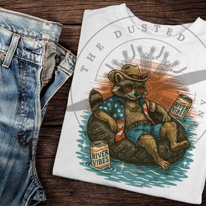 Peut inclure: T-shirt blanc avec un graphique d'un raton laveur portant un bandana rouge, blanc et bleu et des lunettes de soleil, assis dans un flotteur de pneu avec une canette de bière "River Vibes". Le texte "The Dusted" est imprimé en cercle autour du raton laveur.