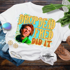 Può includere: T-shirt bianca con la scritta "DROP DEAD FRED DID IT" in lettere arancioni e gialle. La maglietta presenta una grafica di un uomo con i capelli rossi e una camicia verde. Sono visibili anche occhiali da sole, gioielli e sandali.