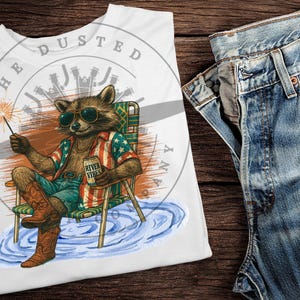 Peut inclure: T-shirt blanc avec un graphique d'un raton laveur portant une chemise rouge, blanche et bleue et tenant une canette de bière qui dit "River Vibes". Le raton laveur est assis sur une chaise en bois avec un pétard dans sa main. Le texte "The Dusted" est écrit en cercle autour du raton laveur.