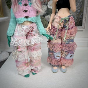 May include: Two dolls wearing handmade doll clothes. One doll is wearing a pink and mint green sweater with floral pants and white lace trim. The other doll is wearing floral patchwork pants with lace trim and blue shoes.