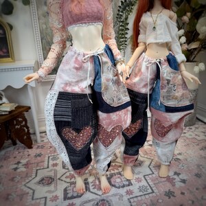 May include: Two dolls wearing patchwork pants with heart-shaped sequin details. The pants feature a mix of denim, patterned fabric, and lace. The dolls are also wearing crop tops. The color palette includes pink, blue, and white.
