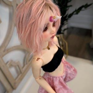 May include: A doll with pink hair and a small unicorn horn. The doll wears a black top and a pink skirt. A gold necklace is around the doll's neck. The doll's eyes are red.