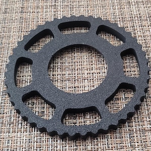 May include: Black plastic gear with eight evenly spaced cutouts around the perimeter. The gear has a textured surface.