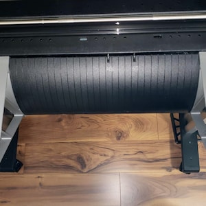 May include: A close-up of a black and silver printer with a roll of paper. The printer is on a wooden floor. The printer has a label that says "142" and "67".
