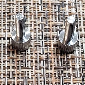 May include: Four silver metal screws with a knurled head. The screws are on a woven brown background.