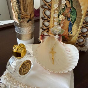 May include: A religious display featuring a candle with a gold and silver design, a framed image of the Virgin Mary, a seashell, a rosary, and a small perfume bottle with a gold cap. The items are arranged on a white cloth.