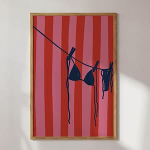 May include: Framed art print showing a navy blue bikini hanging on a washing line against a background of red and pink vertical stripes. The artwork has a minimalist, beachy feel.
