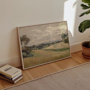 K&ouml;nnte beinhalten: Ein gerahmtes Landschaftsgem&auml;lde in neutralen Farbt&ouml;nen. Das Kunstwerk zeigt ein Feld mit B&auml;umen und sanften H&uuml;geln unter einem bew&ouml;lkten Himmel. Das Gem&auml;lde lehnt an einer wei&szlig;en Wand, neben einem Stapel B&uuml;cher und einer Topfpflanze.