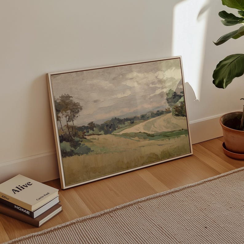 K&ouml;nnte beinhalten: Ein gerahmtes Landschaftsgem&auml;lde in neutralen Farbt&ouml;nen. Das Kunstwerk zeigt ein Feld mit B&auml;umen und sanften H&uuml;geln unter einem bew&ouml;lkten Himmel. Das Gem&auml;lde lehnt an einer wei&szlig;en Wand, neben einem Stapel B&uuml;cher und einer Topfpflanze.