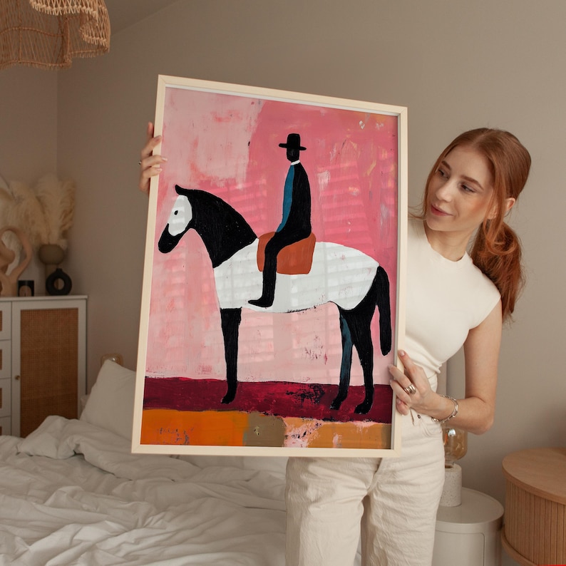 K&ouml;nnte beinhalten: Ein gerahmtes Gem&auml;lde, das ein schwarz-wei&szlig;es Pferd mit einer Person in einem schwarzen Hut und einem blauen Hemd zeigt, die darauf reitet. Der Hintergrund ist ein abstraktes Design in Rosa- und Rott&ouml;nen.