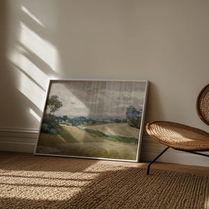 K&ouml;nnte beinhalten: Gerahmtes Landschaftsgem&auml;lde mit sanften H&uuml;geln und B&auml;umen unter einem bew&ouml;lkten Himmel. Das Kunstwerk weist eine ged&auml;mpfte Farbpalette aus Gr&uuml;n-, Gelb- und Graut&ouml;nen mit einem wei&szlig;en Rahmen auf. Das Gem&auml;lde lehnt an einer Wand, neben einem Korbstuhl.