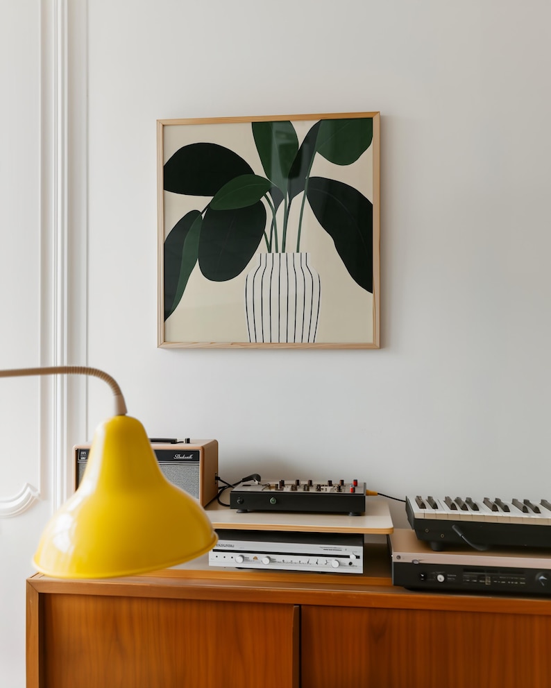 May include: A framed art print featuring a stylized illustration of a plant with large, dark green leaves in a striped vase. The artwork hangs above a wooden cabinet with a yellow lamp, amplifier, and electronic music equipment.