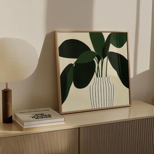 May include: Square framed art print featuring a stylized illustration of a potted plant with large green leaves. The plant is in a white vase with black vertical stripes, set against a cream background. The frame is a light wood color.