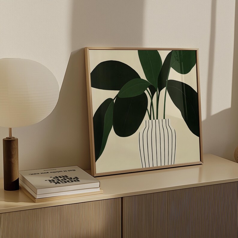 May include: Square framed art print featuring a stylized illustration of a potted plant with large green leaves. The plant is in a white vase with black vertical stripes, set against a cream background. The frame is a light wood color.