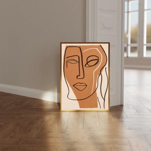 Op de afbeelding: Abstracte lijntekening print van een gezicht met bruine en zwarte lijnen op een beige achtergrond. De print is ingelijst in een gouden lijst.