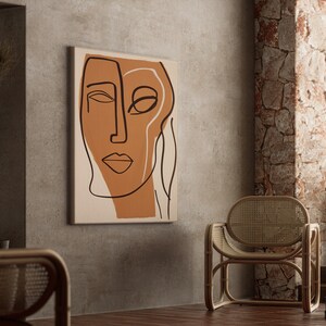 Op de afbeelding: Abstracte lijntekening print van een gezicht met bruine en witte lijnen op een beige achtergrond. De print hangt aan een grijze muur in een kamer met een rieten stoel en een stenen muur.