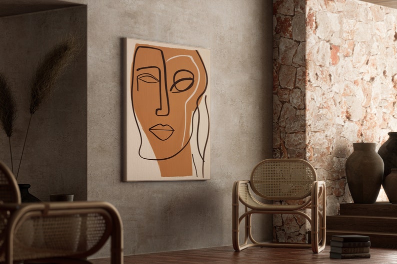 Op de afbeelding: Abstracte lijntekening print van een gezicht met bruine en witte lijnen op een beige achtergrond. De print hangt aan een grijze muur in een kamer met een rieten stoel en een stenen muur.
