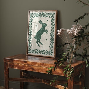 May include: A framed print of a green rabbit jumping in a leafy green frame on a brown wooden table.