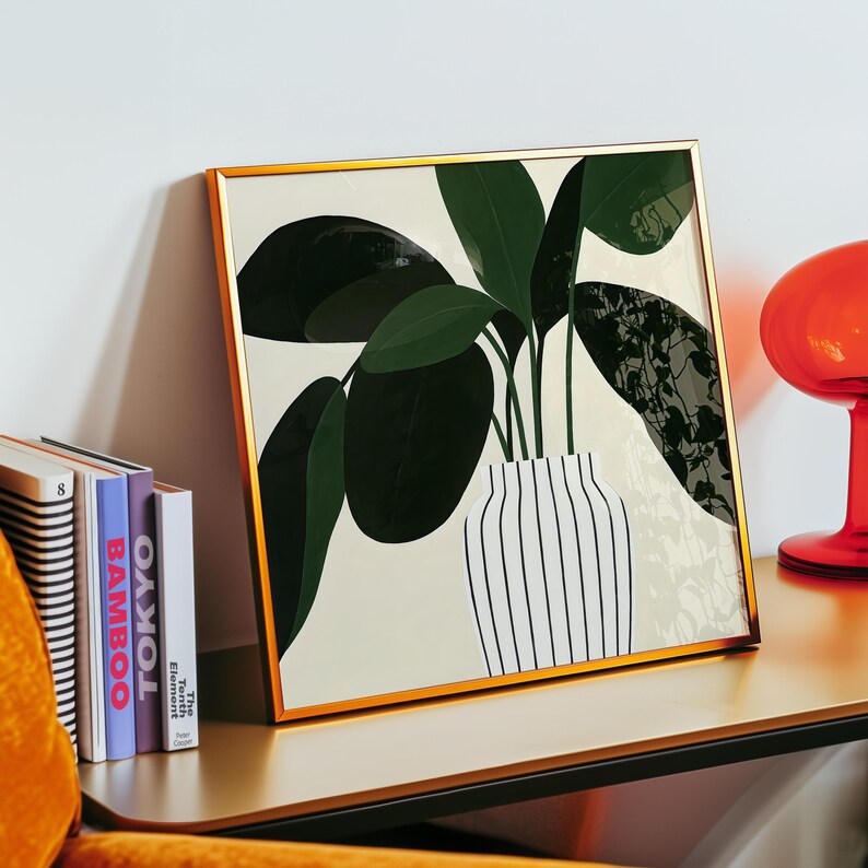May include: A framed art print featuring a stylized illustration of a houseplant in a striped vase. The artwork has a gold frame and is displayed on a gold-colored table. The plant has large, dark green leaves. Books are visible to the left.