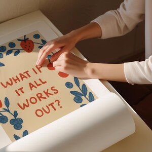 K&ouml;nnte beinhalten: Ein Poster mit dem Text "WHAT IF IT ALL WORKS OUT?" in roten Buchstaben. Das Poster hat einen beigen Hintergrund und ist mit Illustrationen von Erdbeeren, Blaubeeren und Bl&auml;ttern verziert. Das Poster wird von einer Person gehalten.