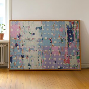 May include: Abstract painting in a wooden frame. The artwork features a blend of colours, including pink, blue, purple, and white, with a pattern of white circles. The painting is displayed against a white wall and a wooden floor.