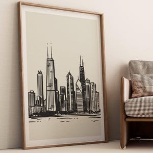 May include: A framed art print with a black and white illustration of a city skyline. The artwork is presented in a light-coloured frame and leans against a wall. The print shows a detailed depiction of various skyscrapers and buildings.