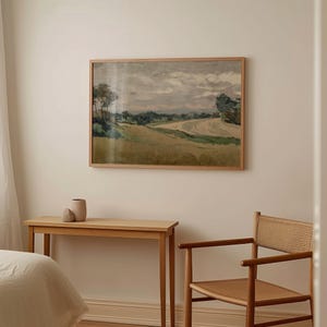 K&ouml;nnte beinhalten: Ein gerahmtes Landschaftsgem&auml;lde mit hellem Holzrahmen h&auml;ngt an einer cremefarbenen Wand. Ein kleiner Holztisch mit einer Vase und ein Stuhl mit gewebter R&uuml;ckenlehne stehen vor der Wand. Das Gem&auml;lde zeigt eine Landschaft mit Feld und B&auml;umen.