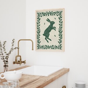 May include: A framed print of a green rabbit silhouette jumping in a green leafy wreath on a cream background.