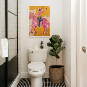 Pu&ograve; includere: Un water bianco con il coperchio chiuso in un bagno con pareti in piastrelle bianche a metro. Un dipinto incorniciato di una persona che cavalca un cavallo rosa &egrave; appeso sopra il water. Una pianta in vaso con foglie verdi si trova sul pavimento accanto al water.