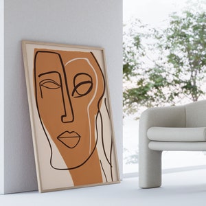Op de afbeelding: Een ingelijst abstract lijntjeskunstprint van een gezicht in bruine en witte kleuren. De print hangt aan een witte muur naast een witte fauteuil.