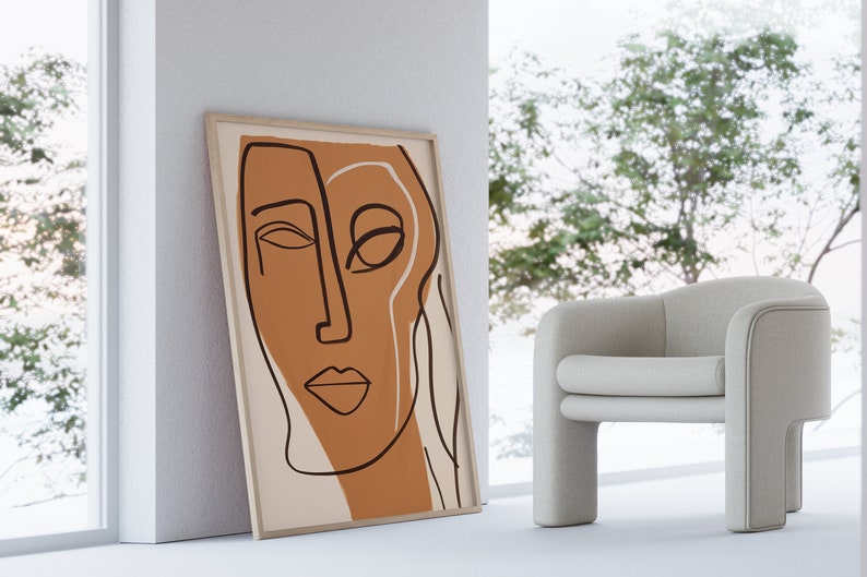 Op de afbeelding: Een ingelijst abstract lijntjeskunstprint van een gezicht in bruine en witte kleuren. De print hangt aan een witte muur naast een witte fauteuil.