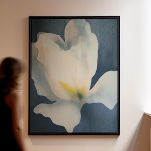 May include: A framed floral art print. The artwork features a large, abstract white flower with yellow and blue accents against a dark blue background. The painting is in a black frame and hangs on a light-colored wall.