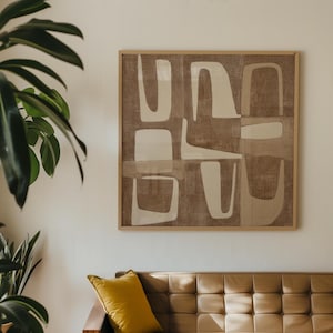 Op de afbeelding: Abstracte wandkunst met een lichtbruine houten lijst. Het kunstwerk heeft een neutraal kleurenpalet met beige en gebroken witte vormen op een getextureerde bruine achtergrond. De kunst hangt boven een bruine leren bank.